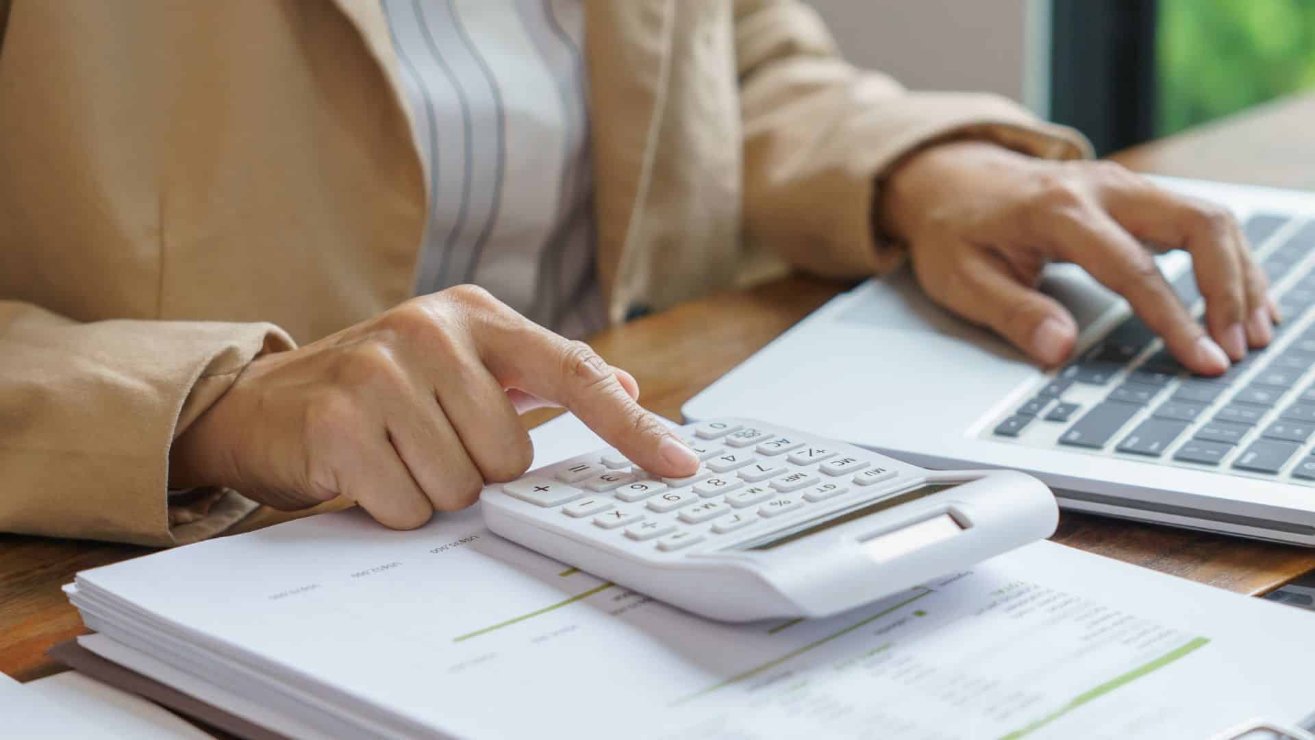 A person uses a calculator while working on a laptop, with documents and charts on the desk.