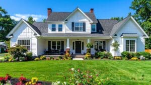 A two-story white house with a covered front porch, gabled roof, and well-maintained lawn and garden under a clear blue sky.