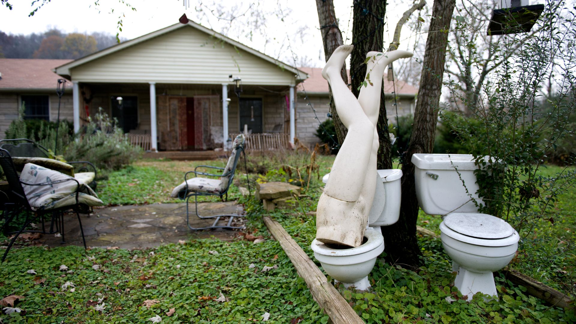 A mannequin torso is propped upside down in a toilet next to another toilet in an overgrown yard in front of a house, with patio chairs nearby.